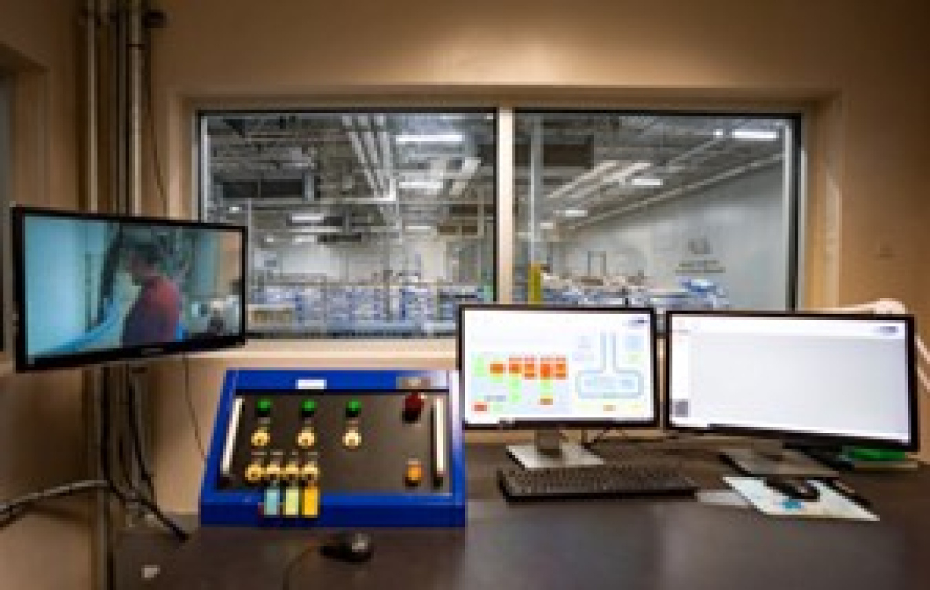 A control room with video displays showing closed-circuit video and readouts from the eBeam process. Next to those is a control panel and behind it all is a window showing the facility.