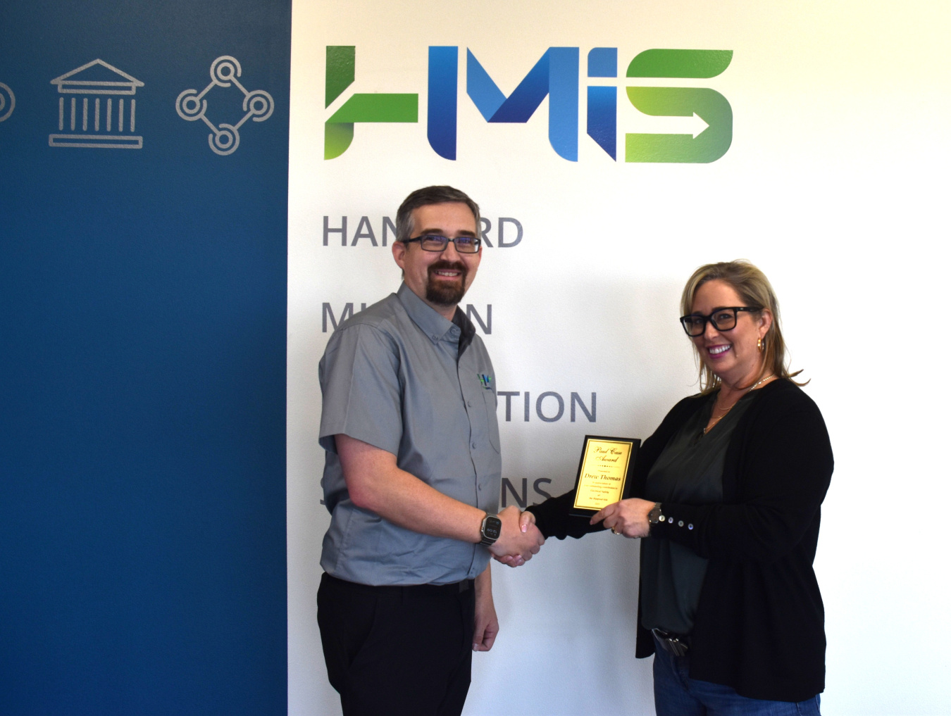 Two professionals shaking hands while smiling for a picture, one holds an award