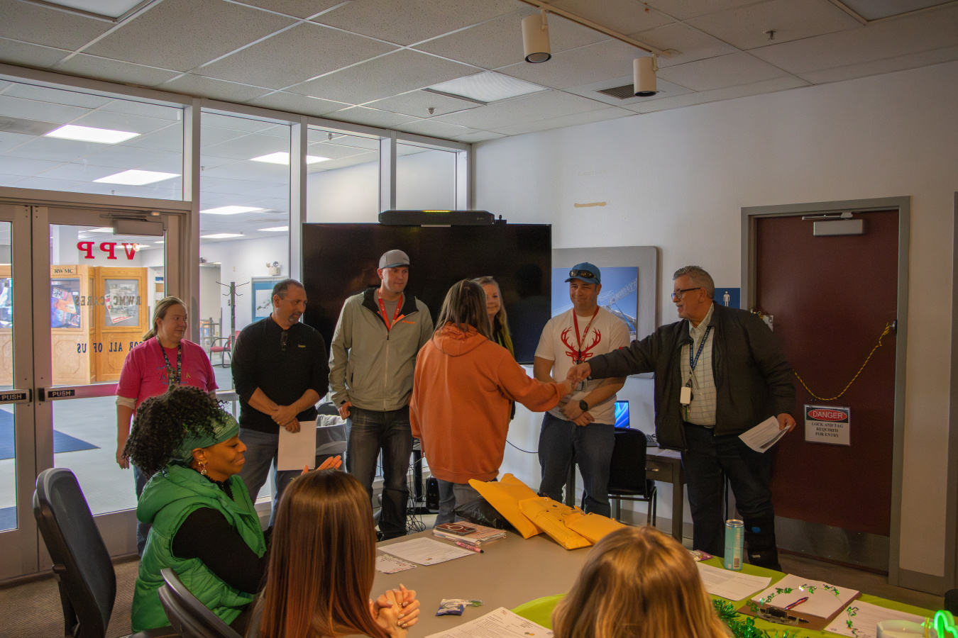 A group of employees in a classroom, some at the front shaking hands with a man while receiving an award