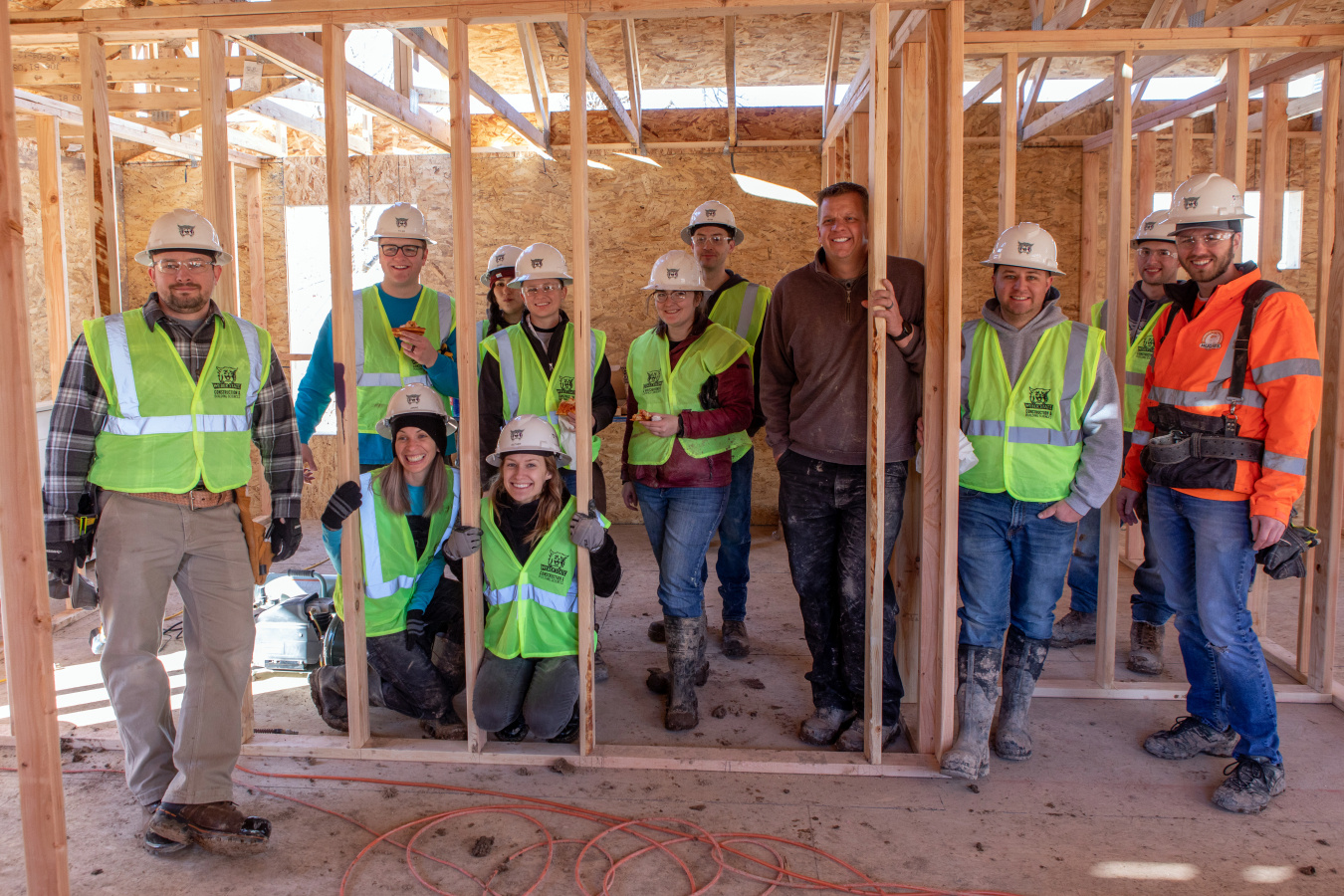 Group of people in construction vests and hard hats standing facing the camera from inside a house under construction.