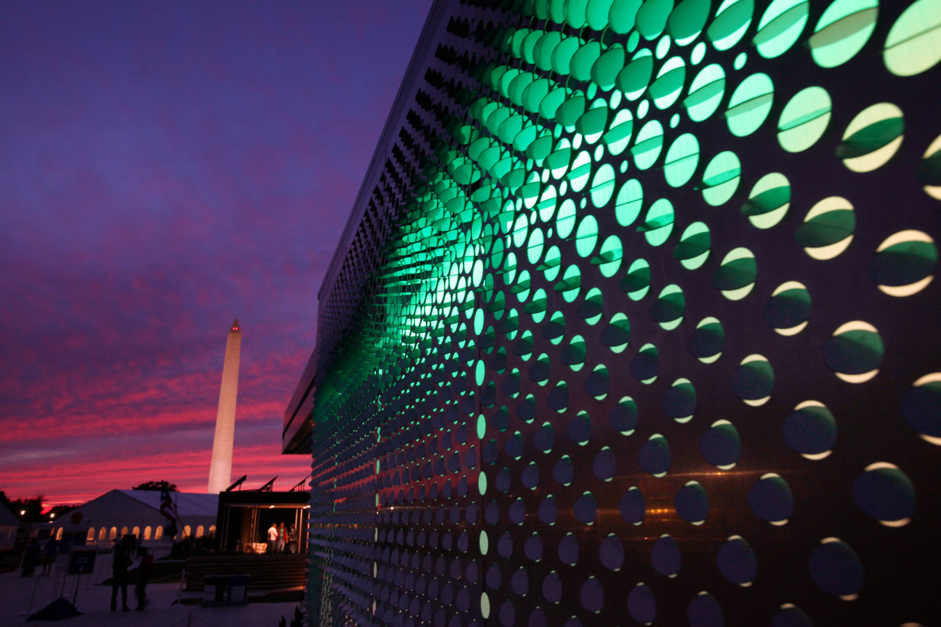 One wall of a Build Challenge competition house, from the outside, showing lights shining inside through the holes in the framework, and you can see the Washington monument in the distance.