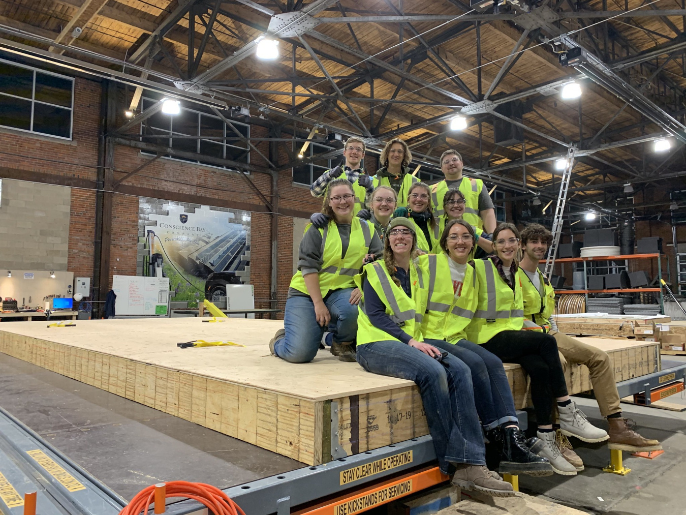 A Build Challenge competition team wearing construction vests and sitting together on top of construction materials, facing the camera, inside a warehouse.