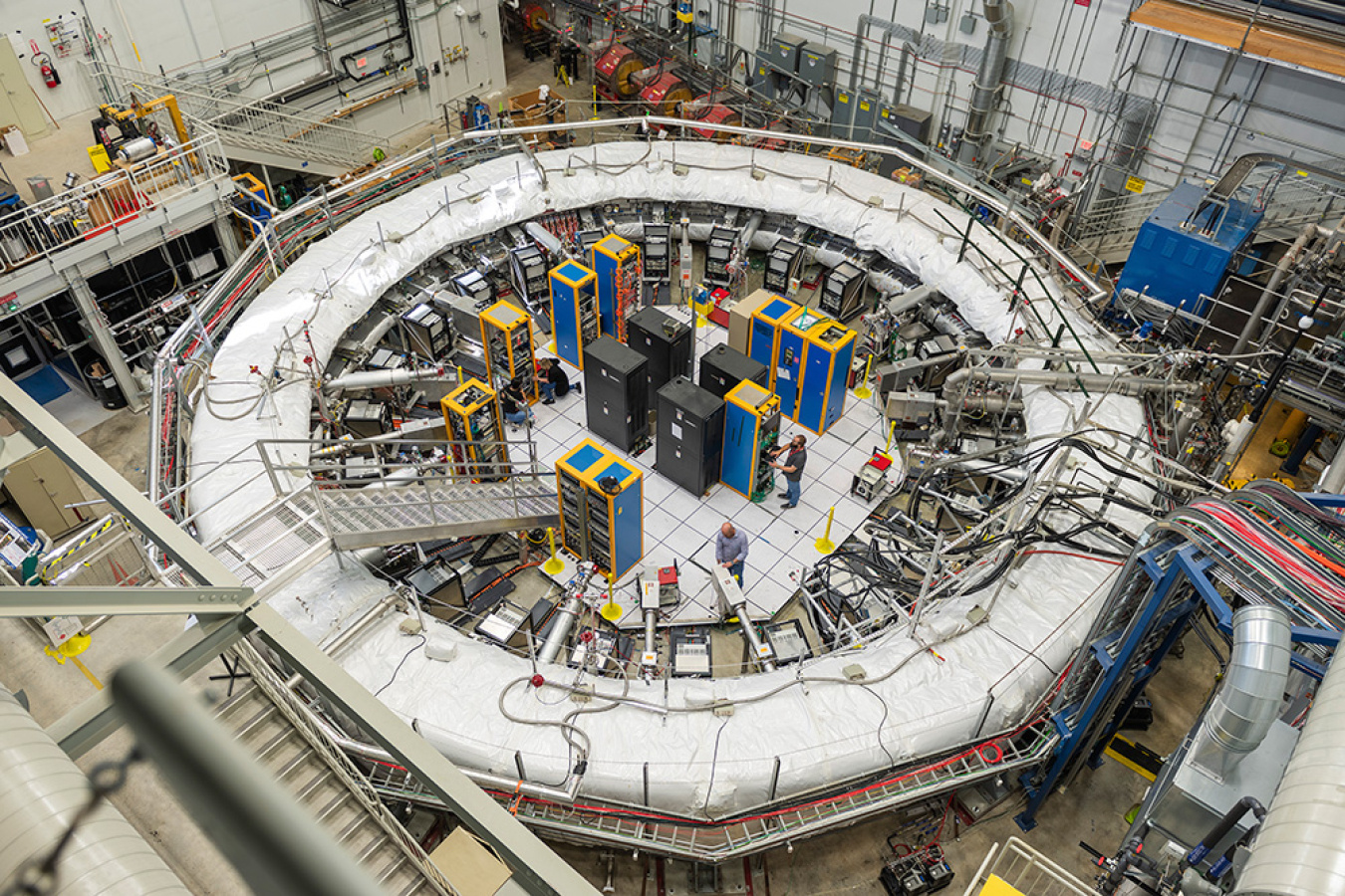 An aerial view of the Muon g-2 experiment, which has a large ring covered in white padding. There are metal arms and tubes coming out of the ring into the middle. There are also computer cabinets and scientists standing inside of the ring. An aerial view of the Muon g-2 experiment, which has a large ring covered in white padding. There are metal arms and tubes coming out of the ring into the middle. There are also computer cabinets and scientists standing inside of the ring. 
