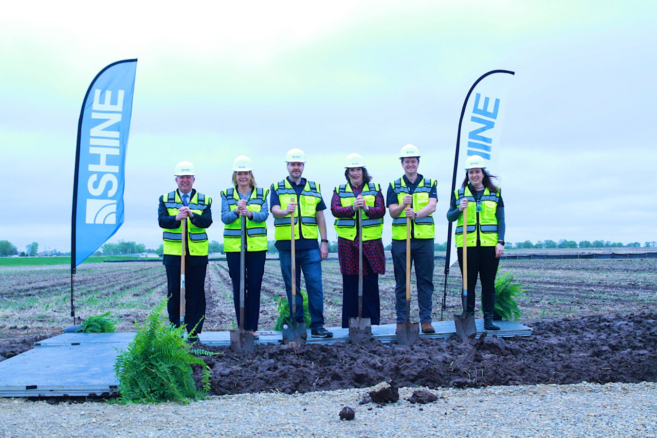 Six people in high-visibility vests and hardhats stand in a field with shovels. Next to them, SHINE-branded banners wave in the wind.