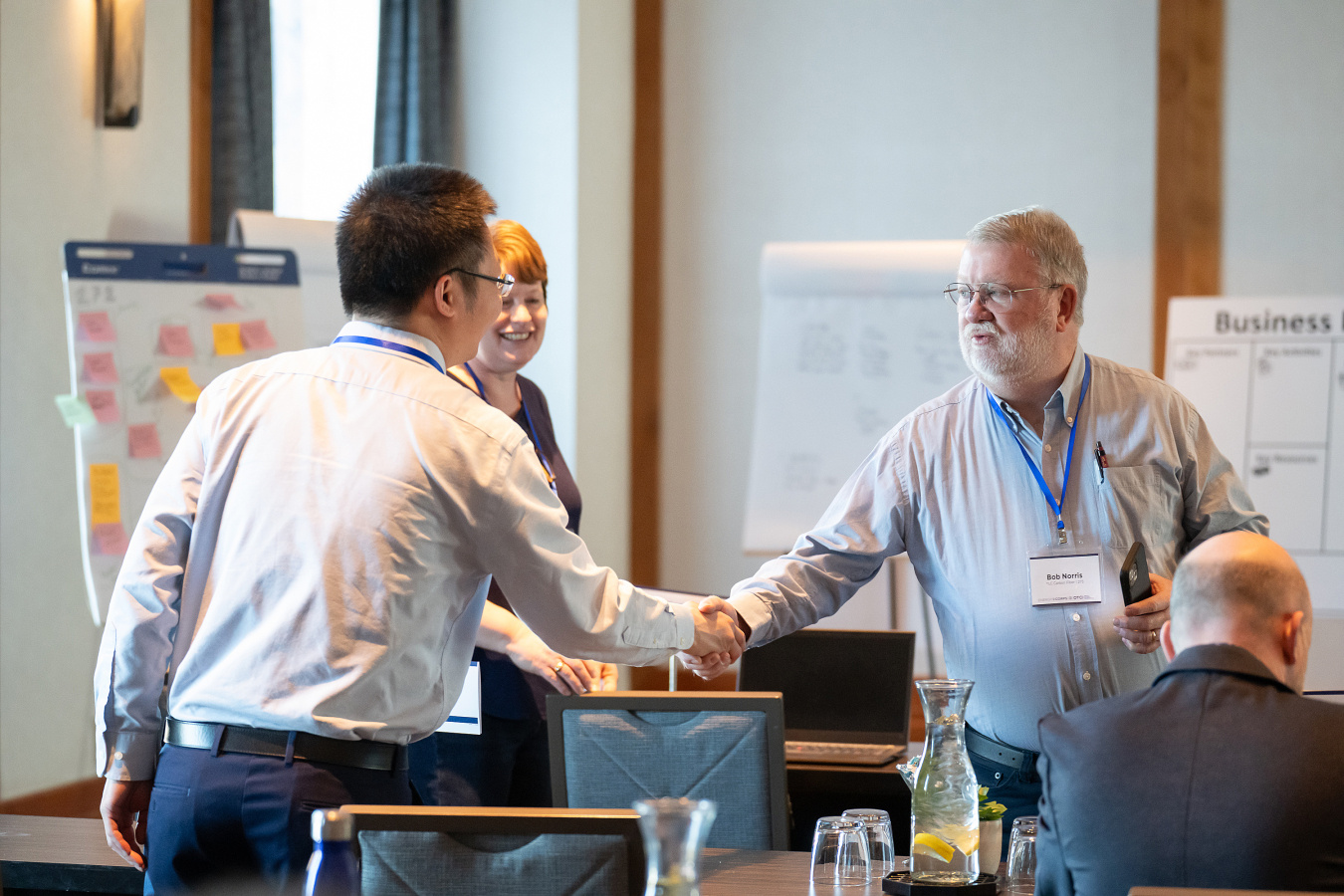 Two men shake hands in introduction with posters and collaborative materials in the background.  
