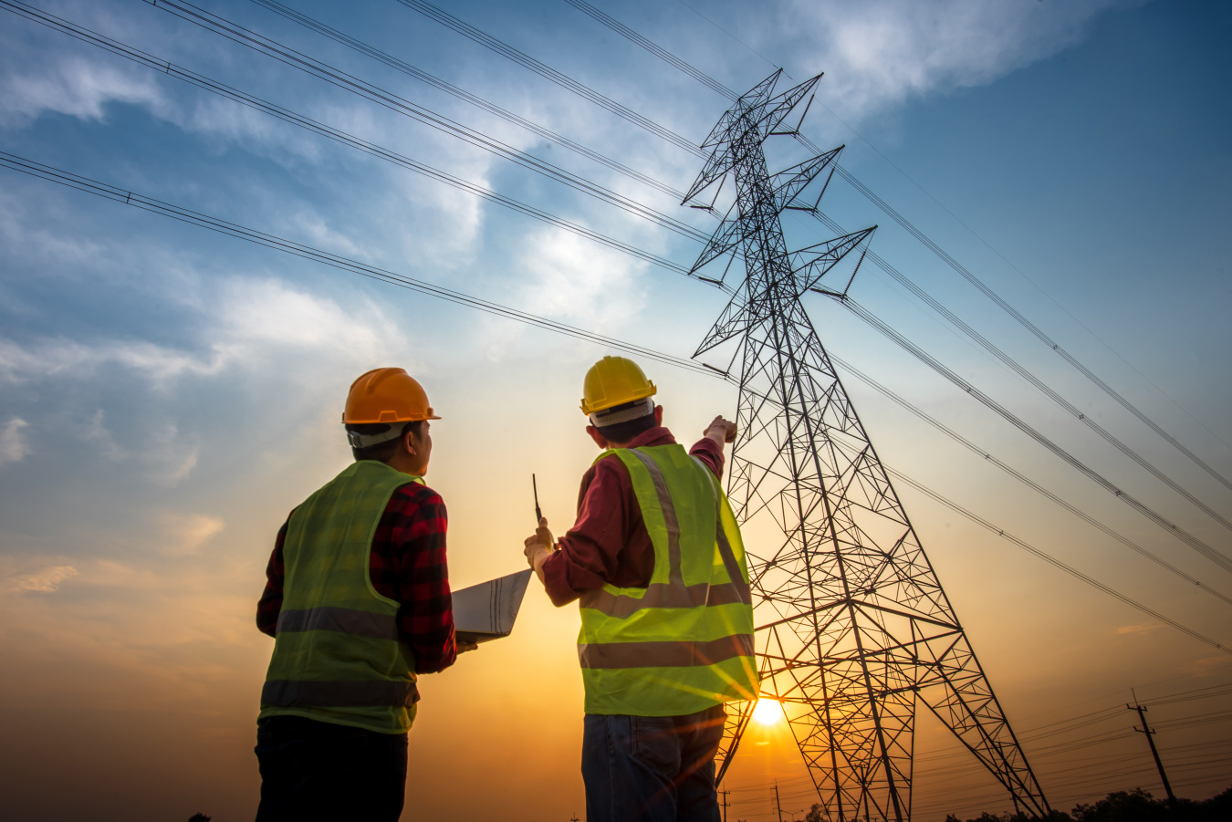 Two workers with a laptop point toward a transmission line tower. 