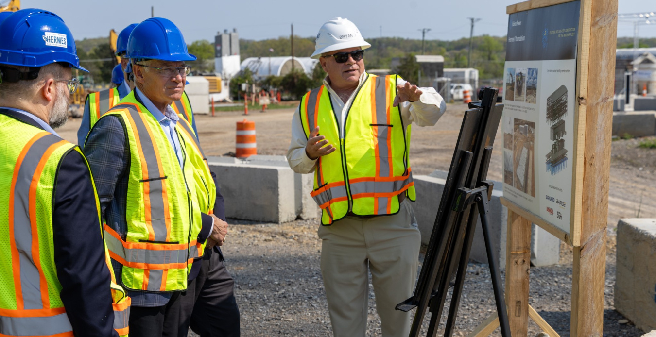 People in reflective vest and hard hats gather around a display and listen to a speaker.