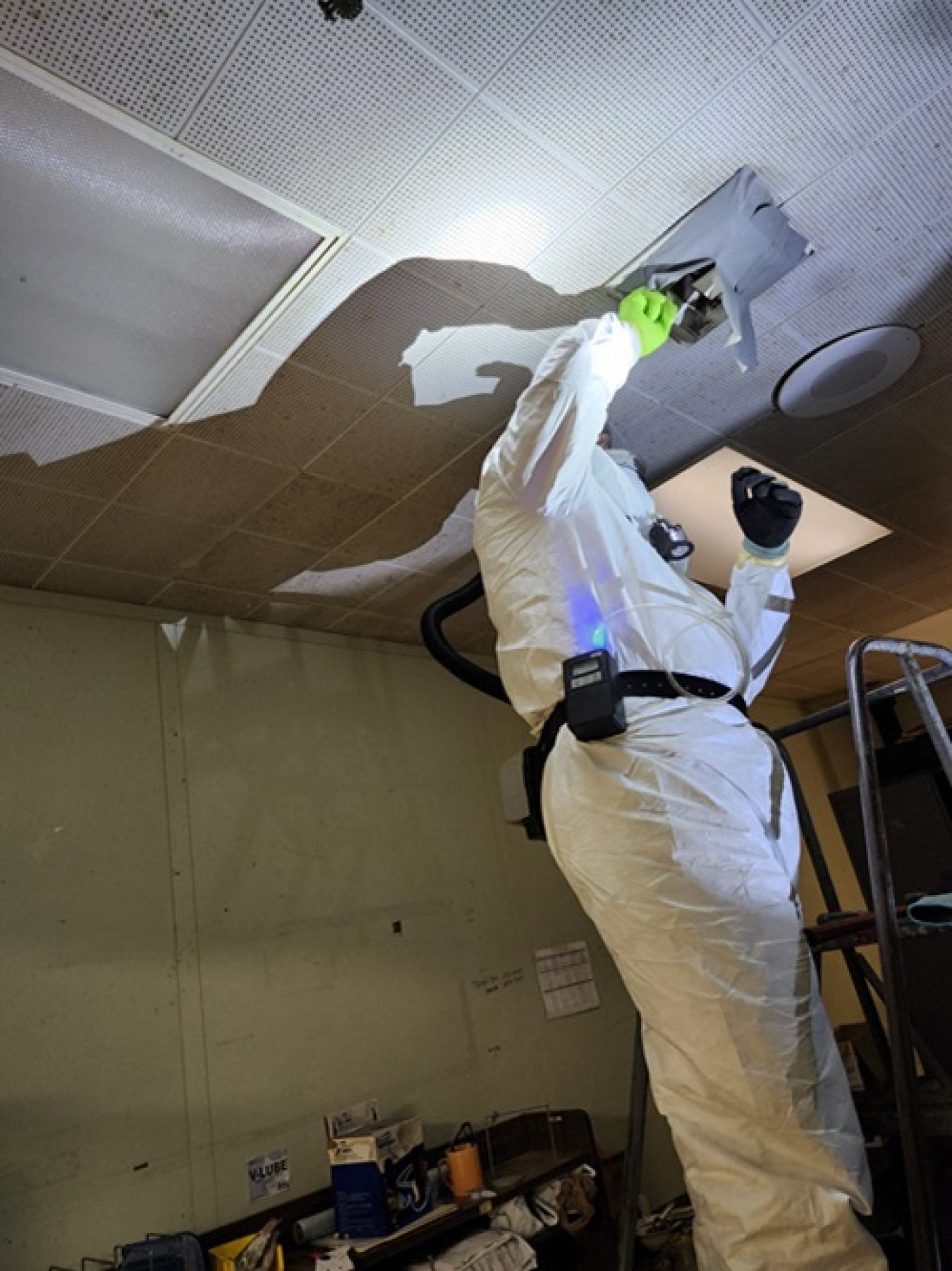 An employee in a white hazmat suit standing on a ladder and reaching into the ceiling
