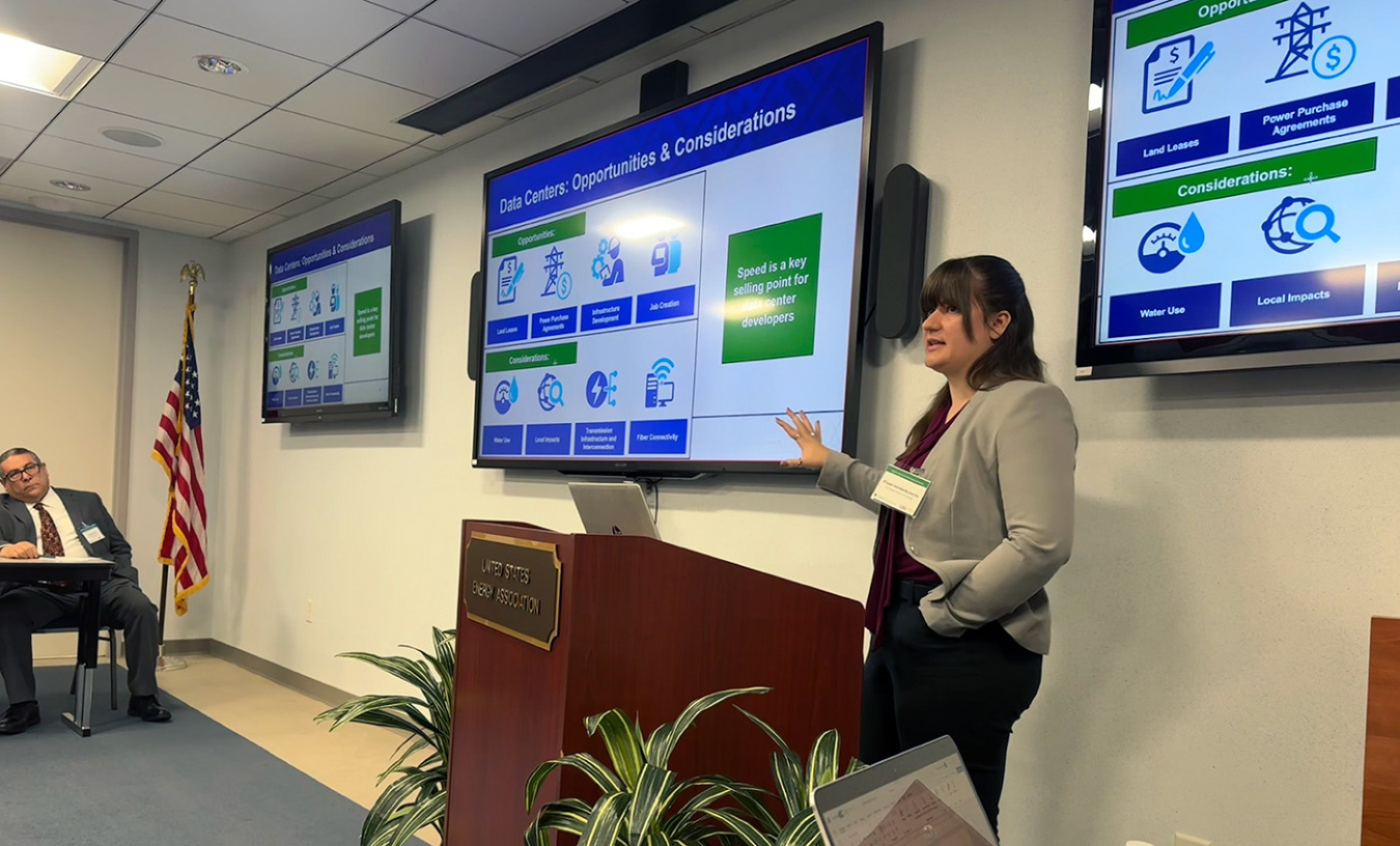 A person wearing a blazer speaking at a podium with a slide showing on televisions behind her.