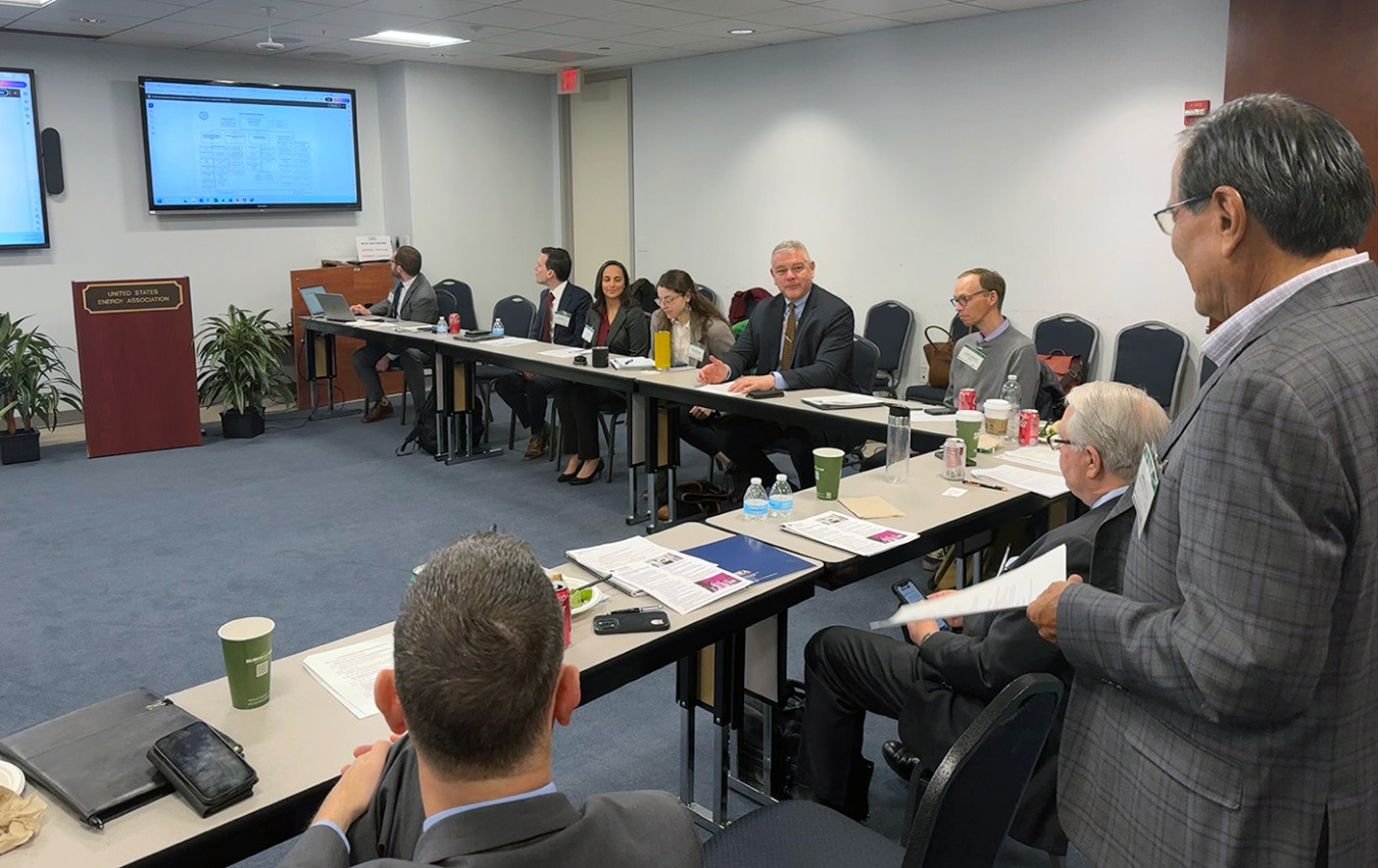 A person wearing a suit jacket, photographed from behind, standing up in a room of about eight people sitting at a large L-shaped desk.