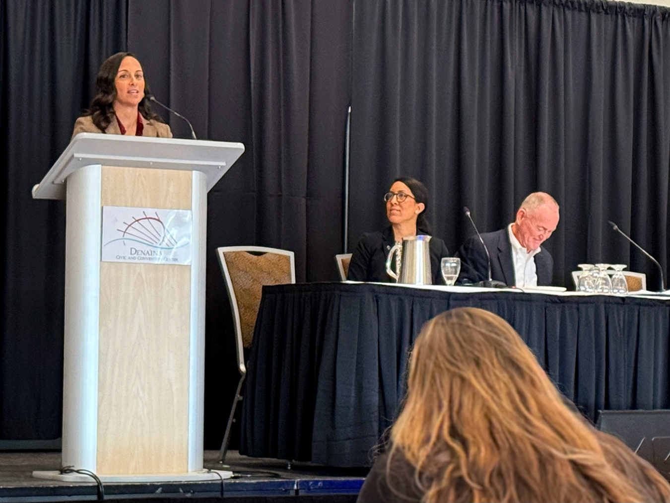 A person on a stage speaking at a podium while two others sitting at a panel table look on.