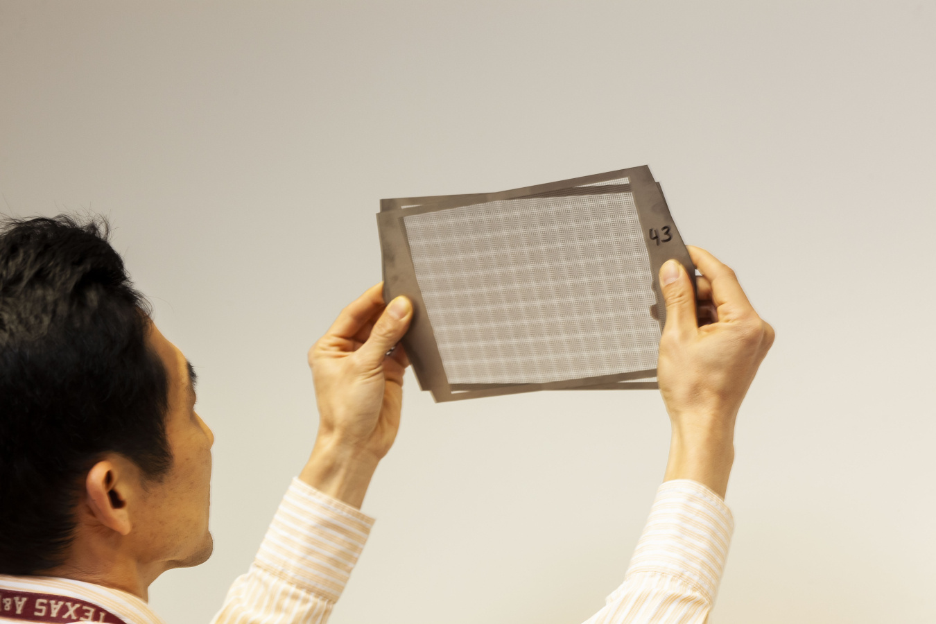 A view over the shoulder of a man holding two overlapping metal sheets over his eye level. The silver metal sheets each have a narrow frame that around a rectangular area perforated with many small holes. The holes in the two sheets line up to create a moire pattern.