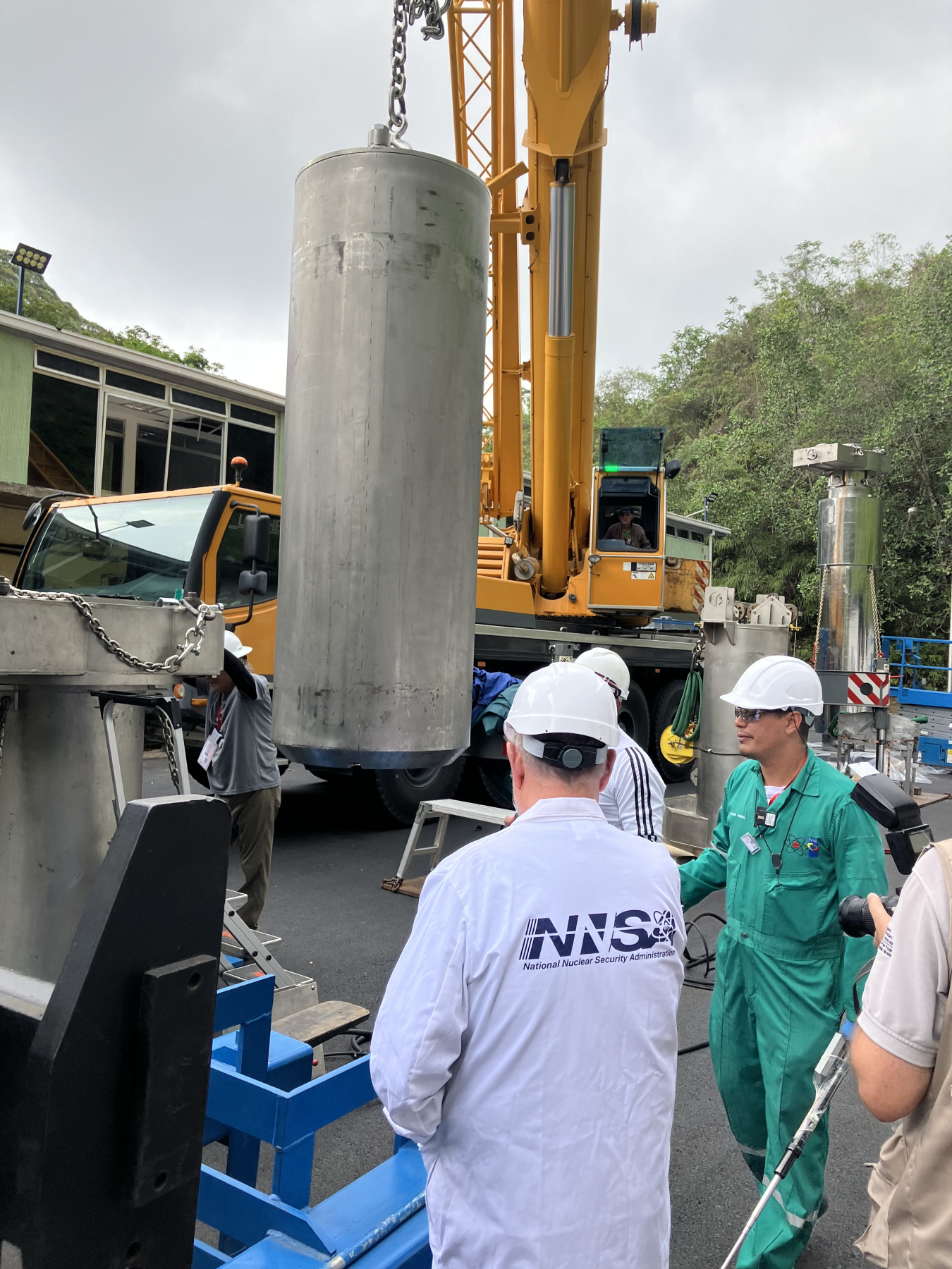 A man wearing an NNSA coat and a hard hat watches as a large cylinder is moved with a crane.