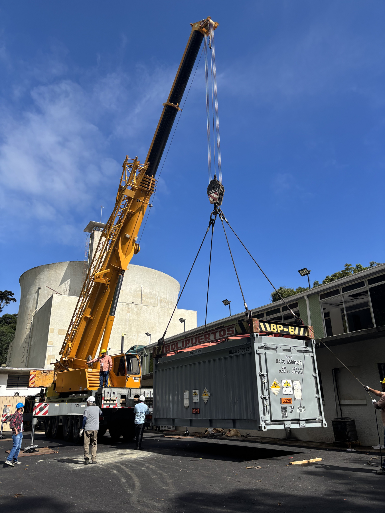 A crane holds a container in the air. A nuclear cooling tower looms in the background.