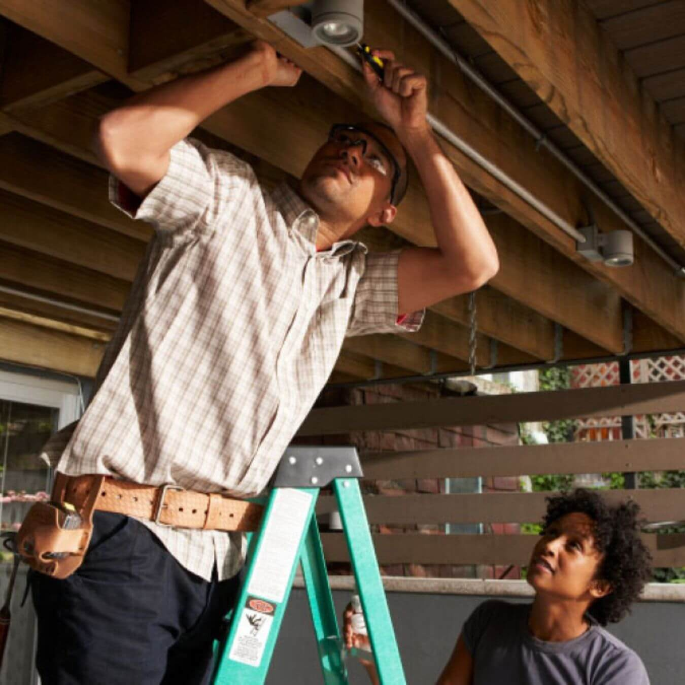 A man on a ladder performs work on lighting overhead while a woman holds the ladder steady.