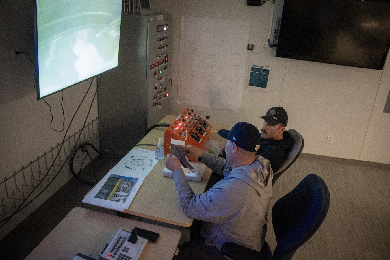 Two men at a control panel desk with a screen on the wall in front of them