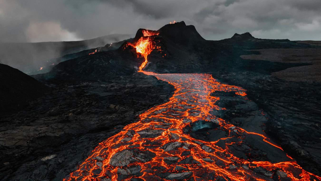 Photo of a small eruption with lava flowing away from it, against a dark rocky landscape with dark clouds in the sky.