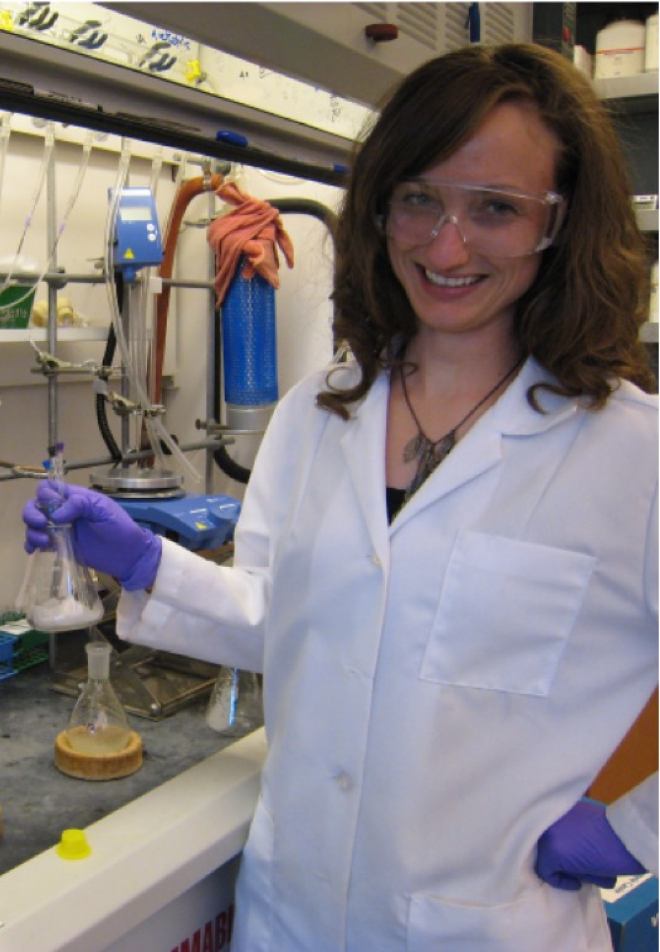 H2O Director Nichole Fitzgerald holding a beaker while wearing a lab coat and safety goggles at her grad school