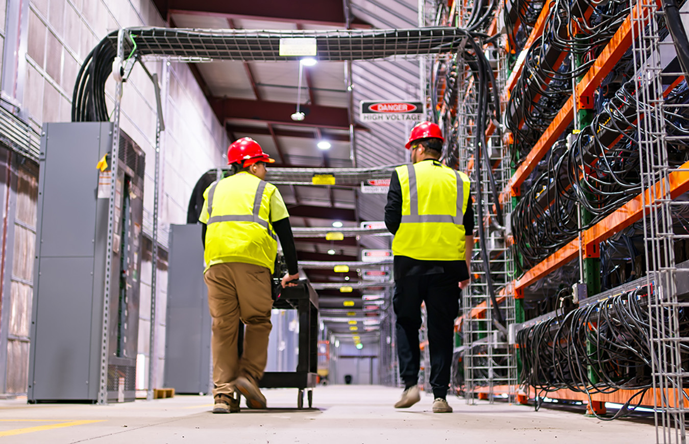 Two technicians wearing PPE walking past racks of equipment in a data center