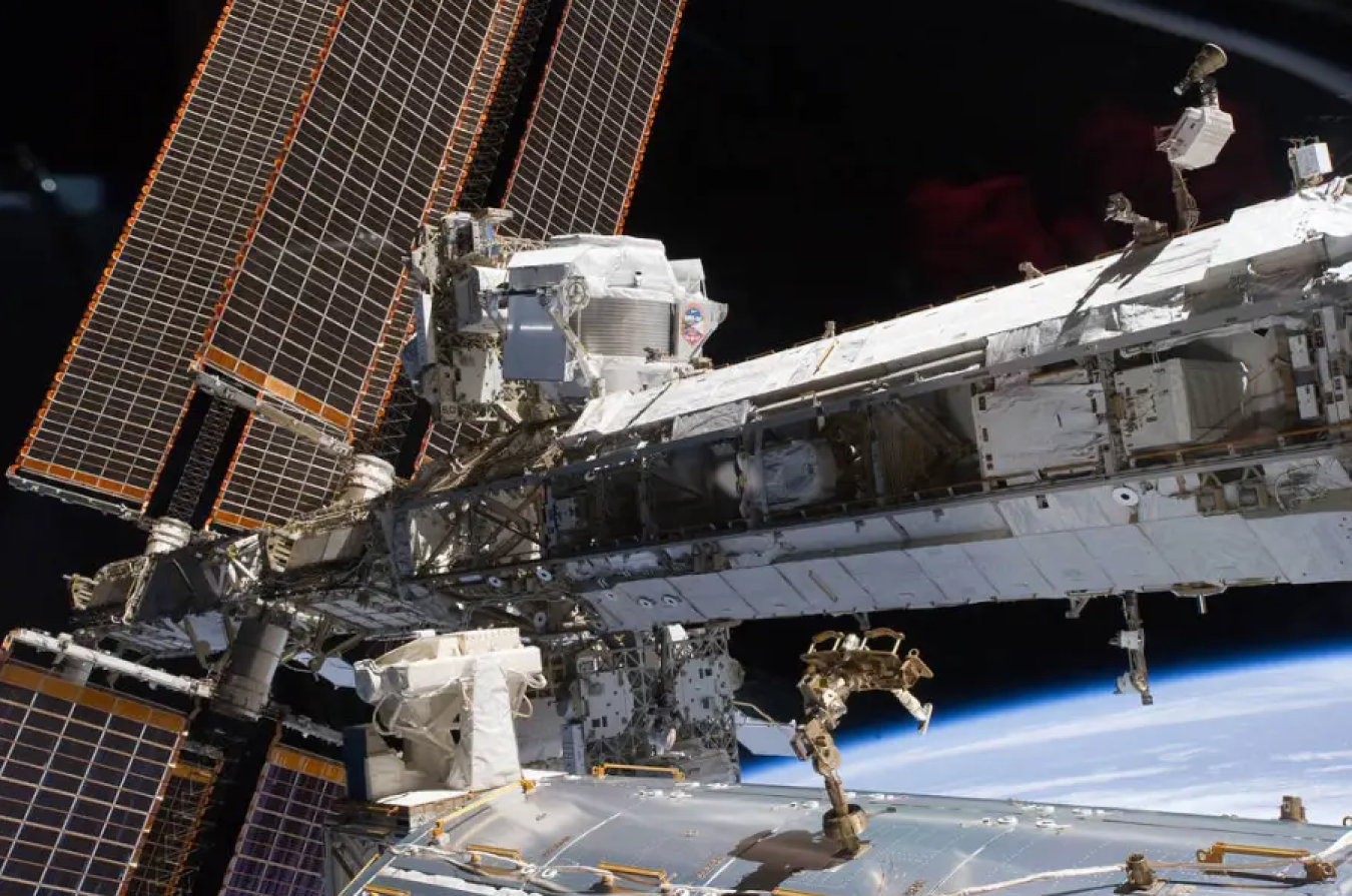 The long tube of the International Space Station runs from left to right across the photo. On the left side, further away from the viewer, large solar panels run from top to bottom. There is a white box-shaped machine just in front of the solar panels, which is the AMS-02. The blackness of space and Earth’s horizon are in the background.