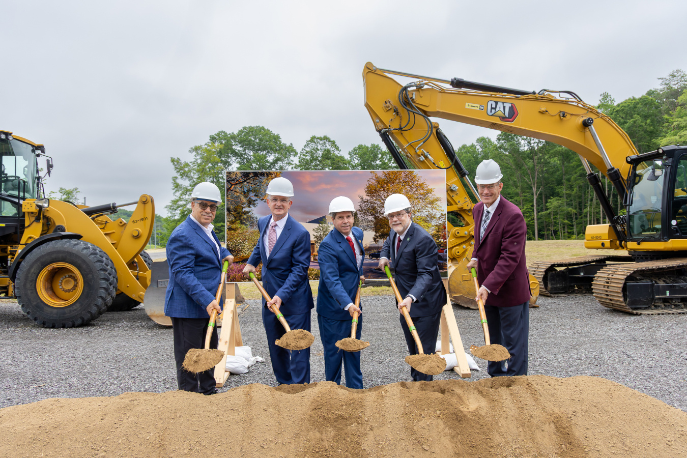 Five men in suits and hard hats shovel dirt from a pile in a pose for the camera. Behind them is earthmoving equipment and a poster showing what the building will look like.