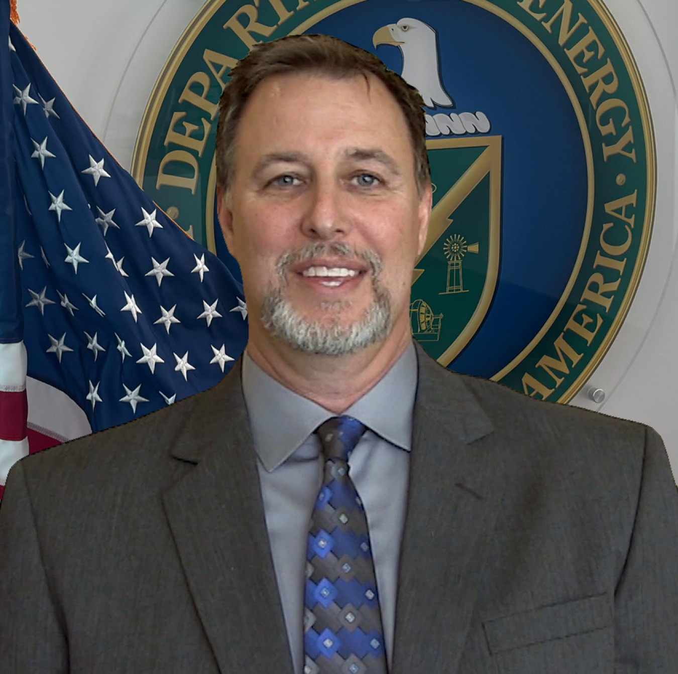 Headshot of a man in a suit in front of the DOE seal and American flag