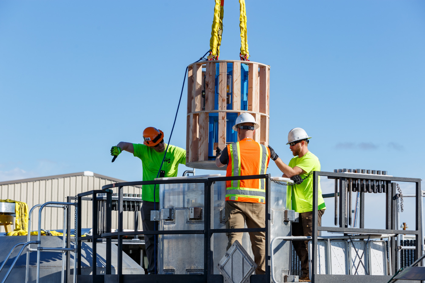 Three men on an elevated platform or truck are dressed in high-visibility vests and hardhats. They are helping guide a crane-held nuclear material transportation cask into place. One of the men is giving a thumbs-down signal.