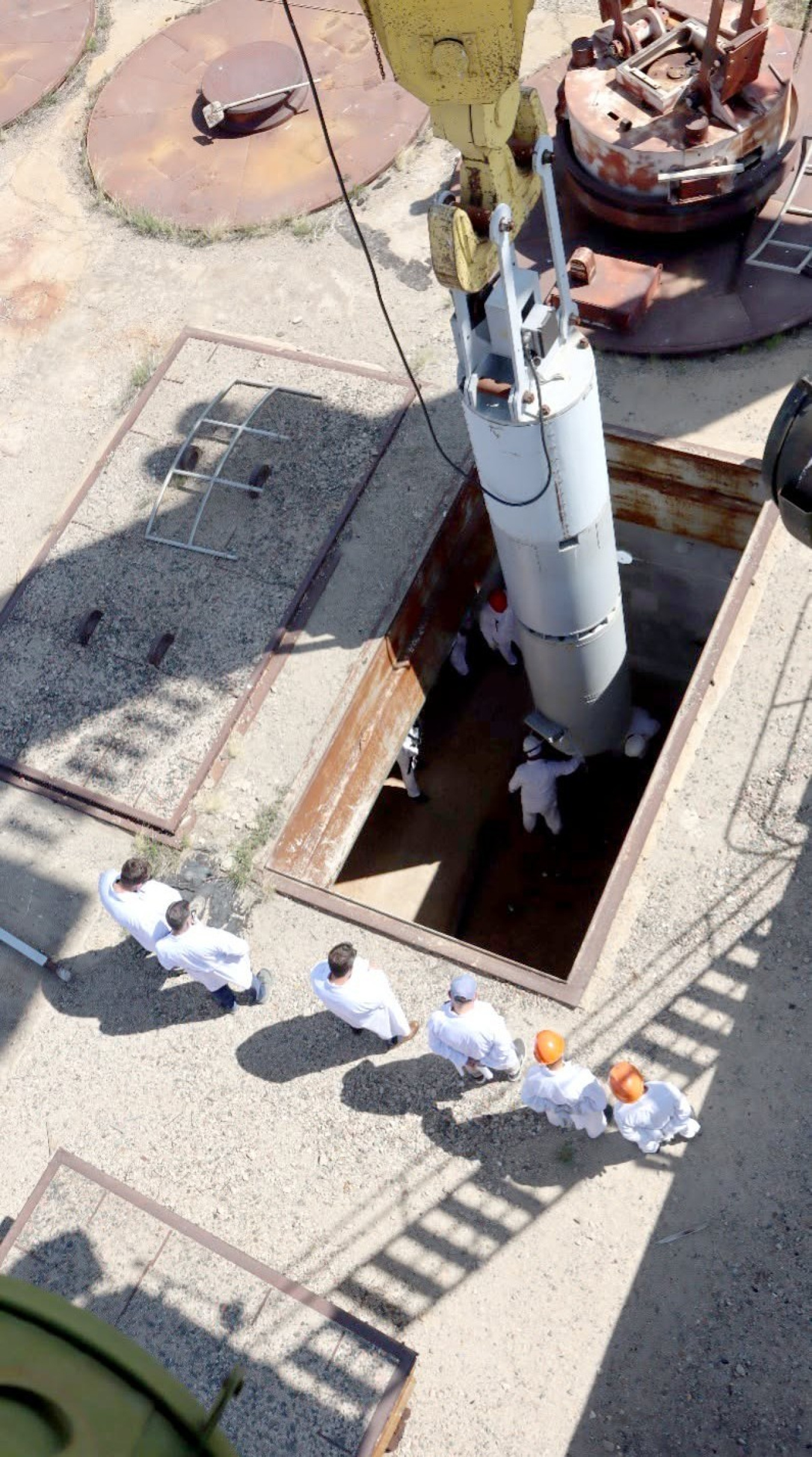 In a photo shot from above, six people dressed in white coats -- some in hardhats, some not -- watch as a long tube is lowered into the ground by a crane.