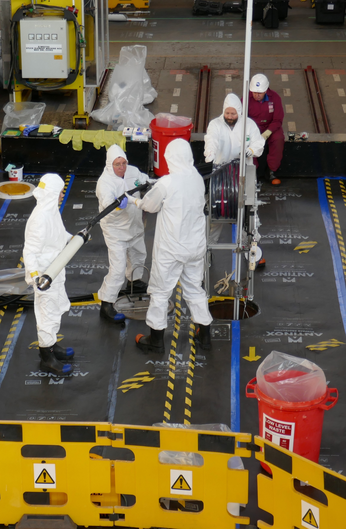 Four people dressed in white, protective "bunny suits" hold a large hose-like piece of equipment in a nuclear facility. 