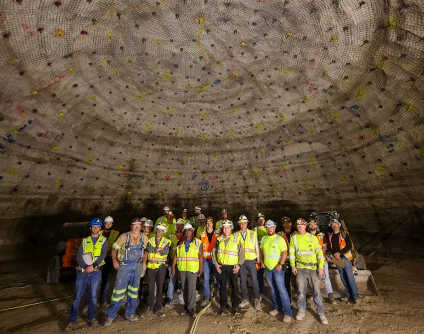 A group of workers wearing high-visibility clothes and hardhats pose for a photo in a large underground chamber.