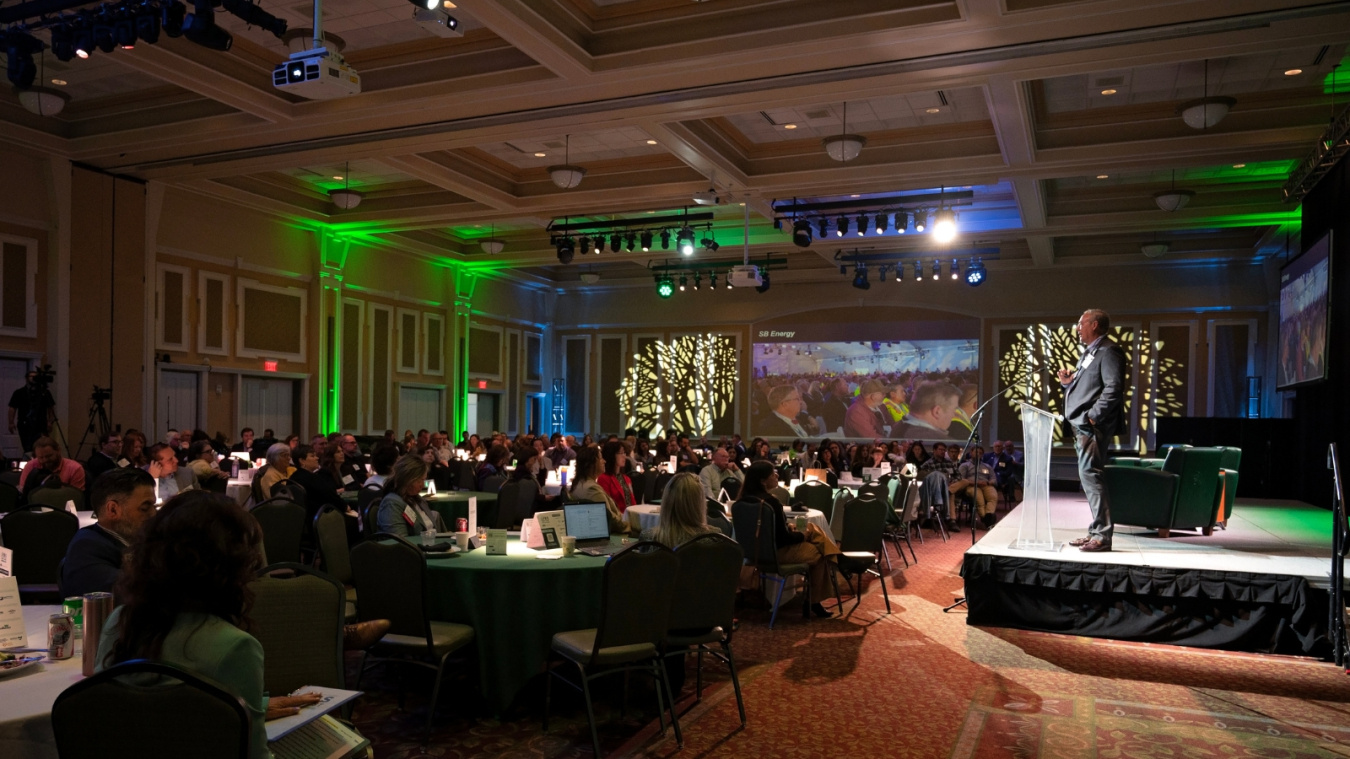 EM leadership on stage behind a podium delivering a speech to a crowd