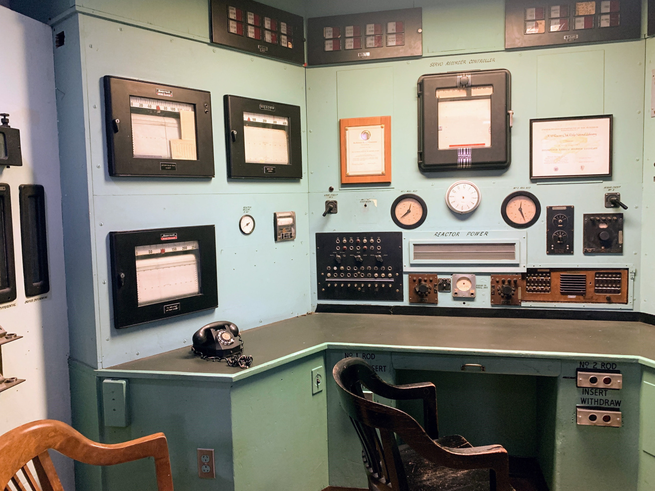 An empty desk in a corner with clocks, control panels and switches on the wall.