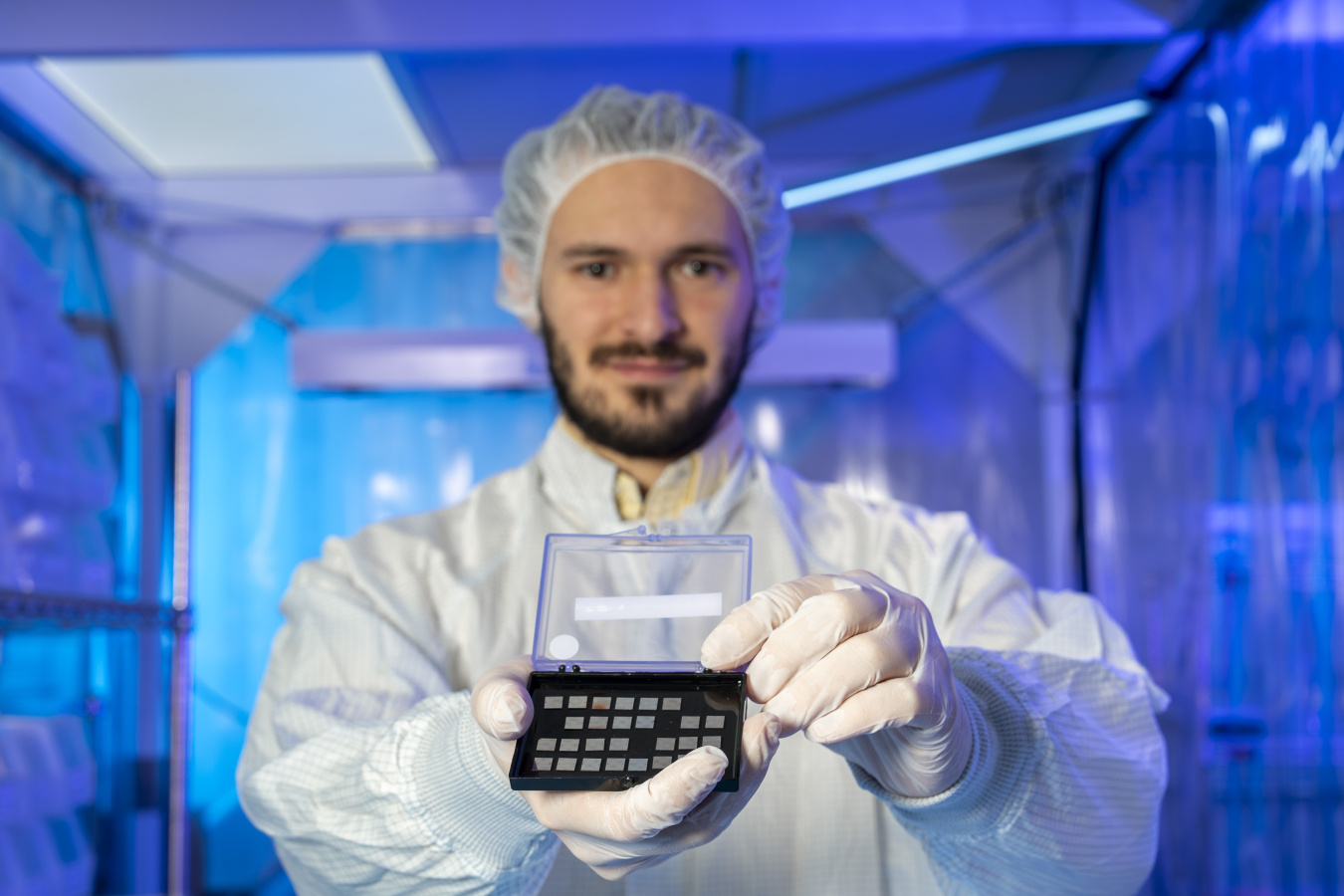 Francesco Crisa (a man in a full clean-room suit) holds out a small plastic box with a series of computer chips in it. Behind him is various clean-room scientific equipment. 