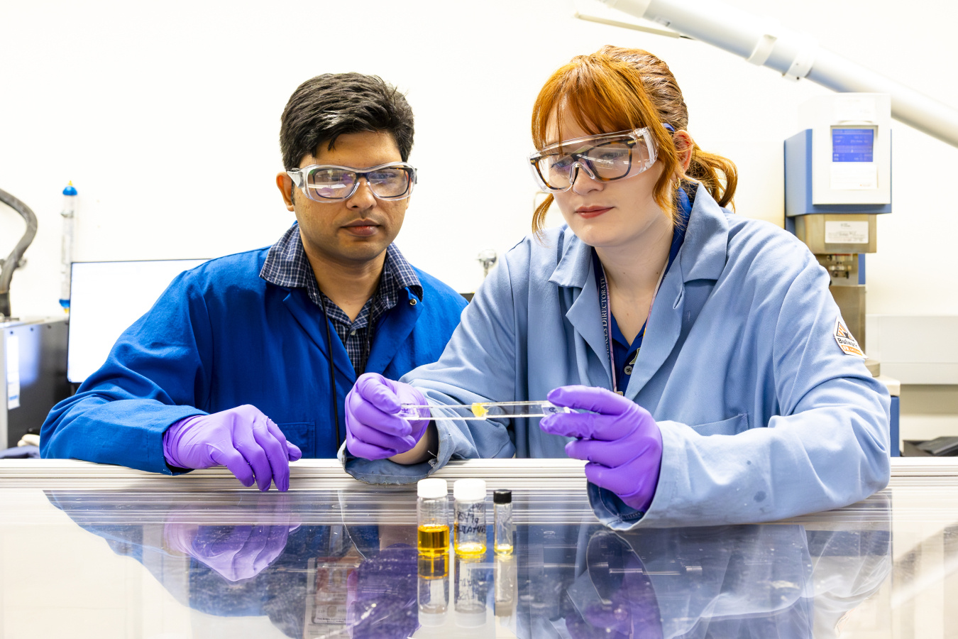 Mary Danielson, who is wearing a lab coat, goggles, and gloves, is holding two pieces of glass attached together with the yellow adhesive. She is leaning on a reflective lab bench with samples of the adhesive in small glass bottles in front of her. Anisur Rahman (who is also in lab equipment) is standing next to her, watching. 