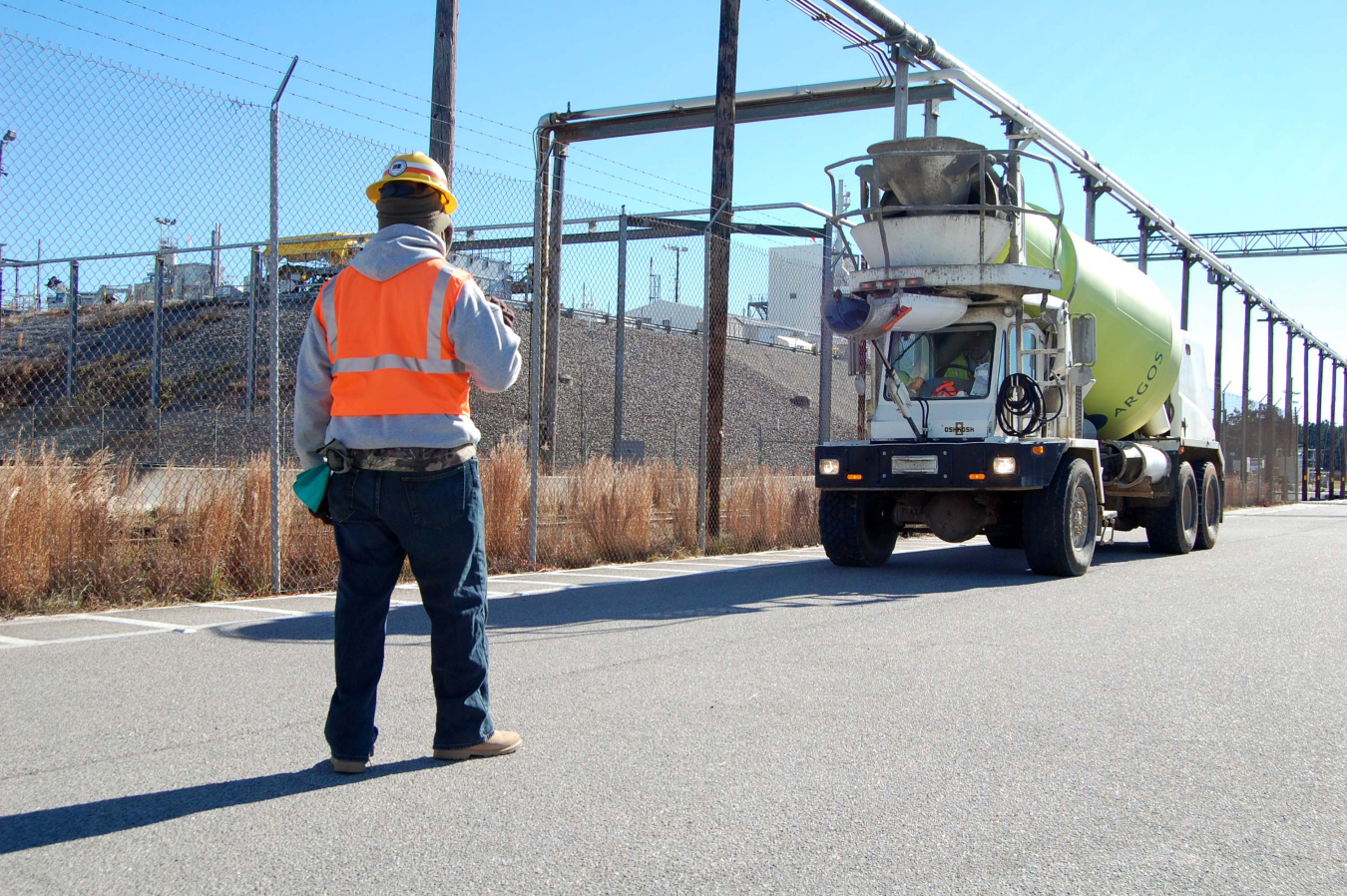 Person wearing an orange safety vest stands with his back toward the camera, next to a security fence and in front of an approaching truck.