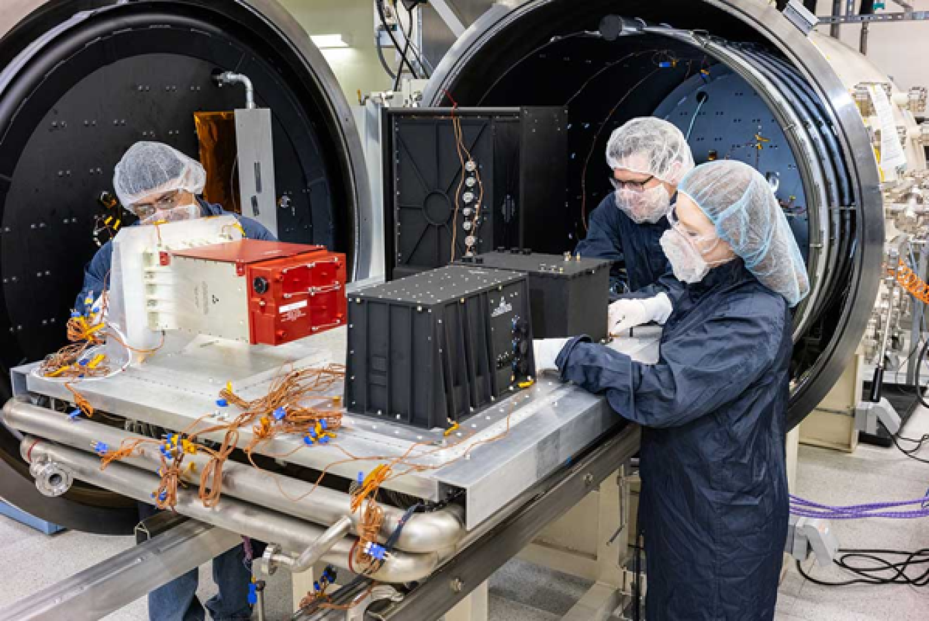 Three people wearing hairnets, masks, and protective clothing, work on a piece of electronic equipment in a laboratory setting.