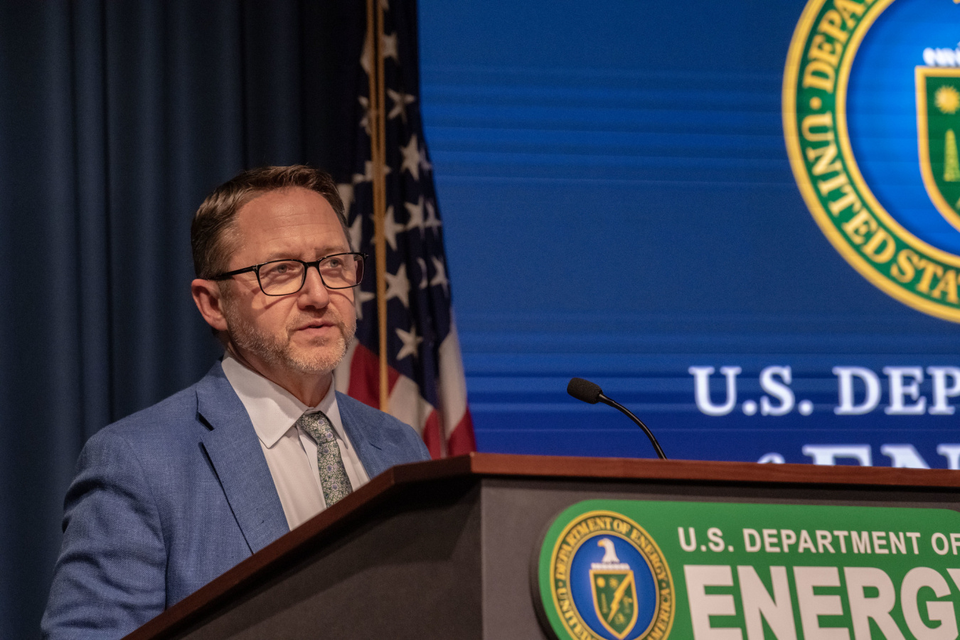  A man stands at a podium on-stage with the U.S. Department of Energy seal displayed. 