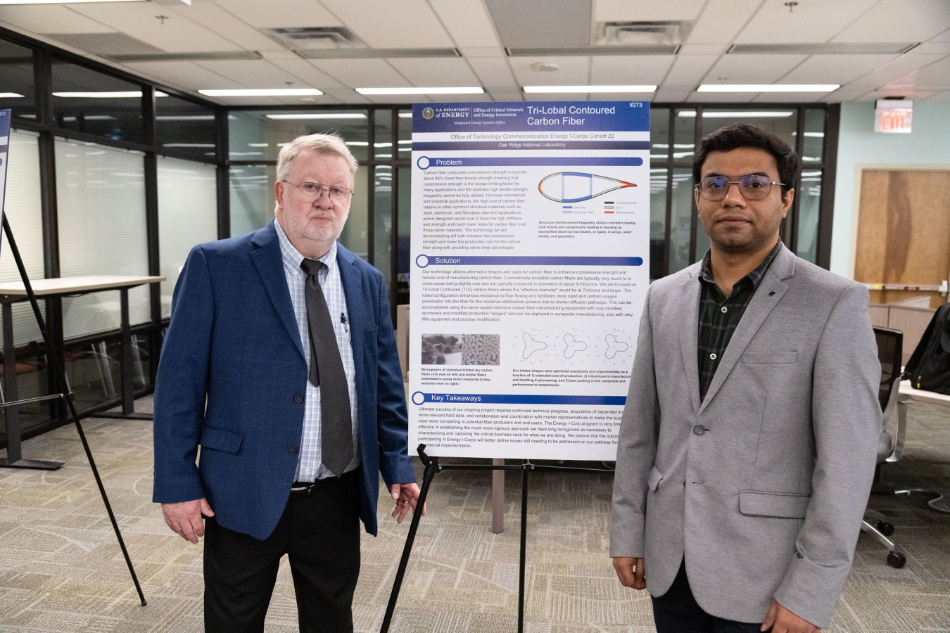 Two men in business suits stand in front of a poster display. 