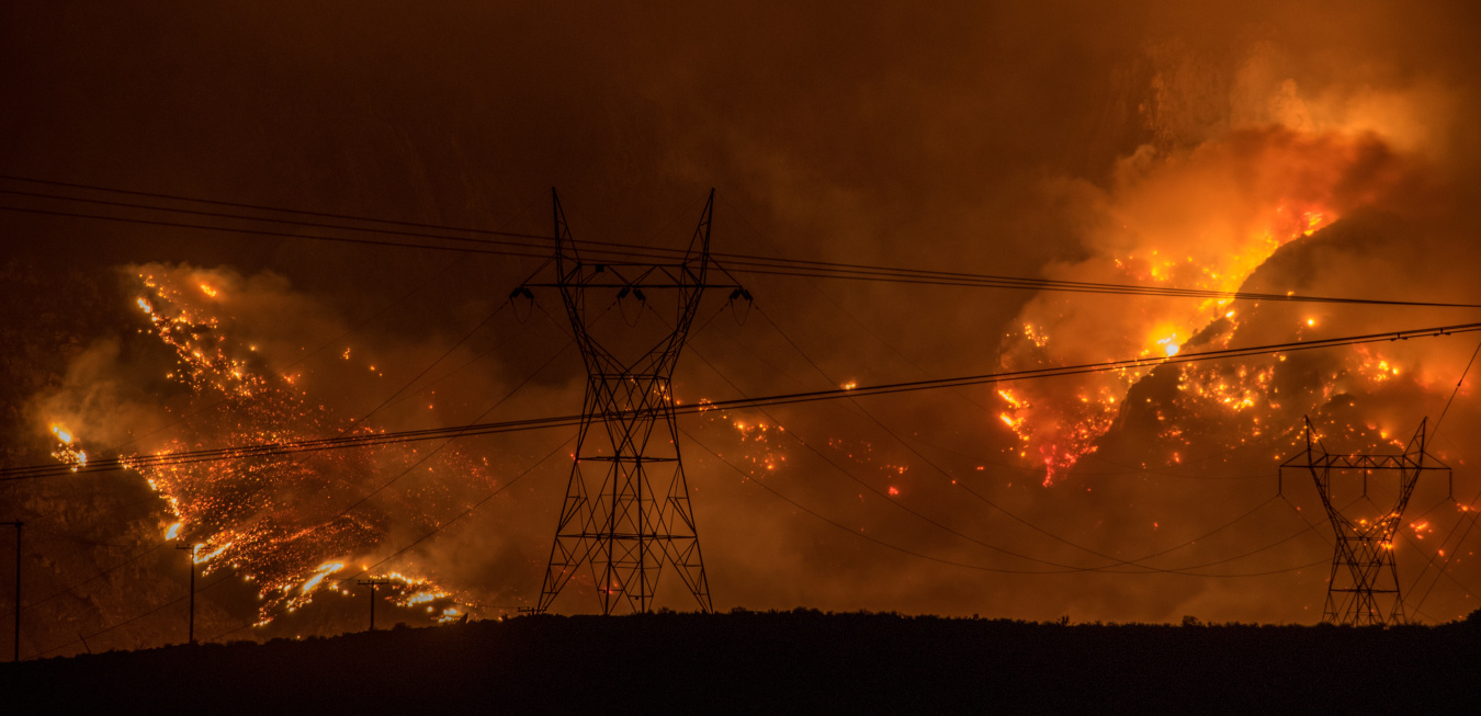 Transmission towers and power lines silhouetted against a large wildfire burning across a hillside at night.