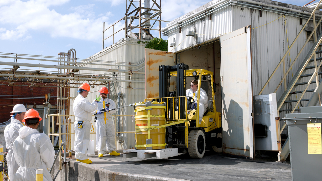 Employees in hazmat suits use construction vehicles to move yellow containers