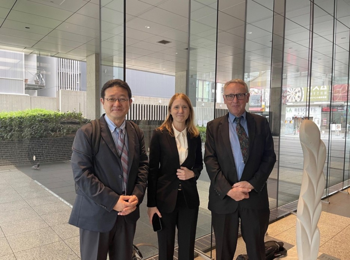 Three United States and Japanese Embassy Officials pose for a photo by a wall of windows