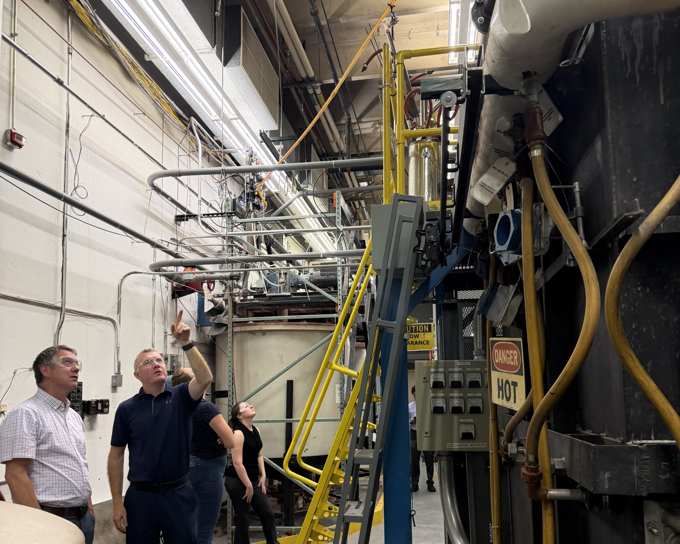 Employees in safety goggles in a laboratory building stand beside a melter and look up at it