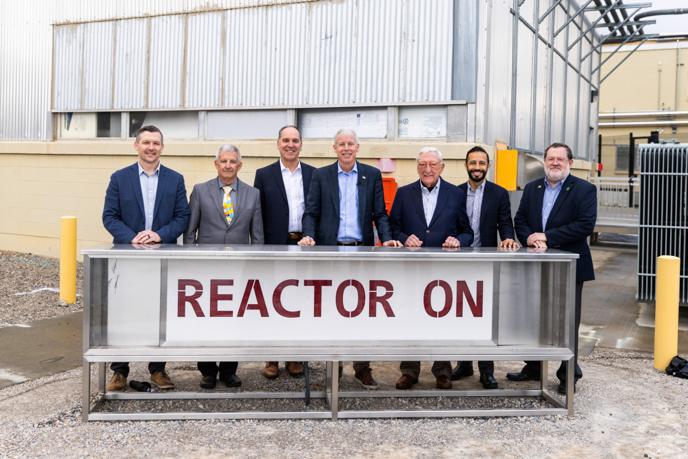 A group photo of DOE leadership standing in front of a sign that says "Reactor On" 