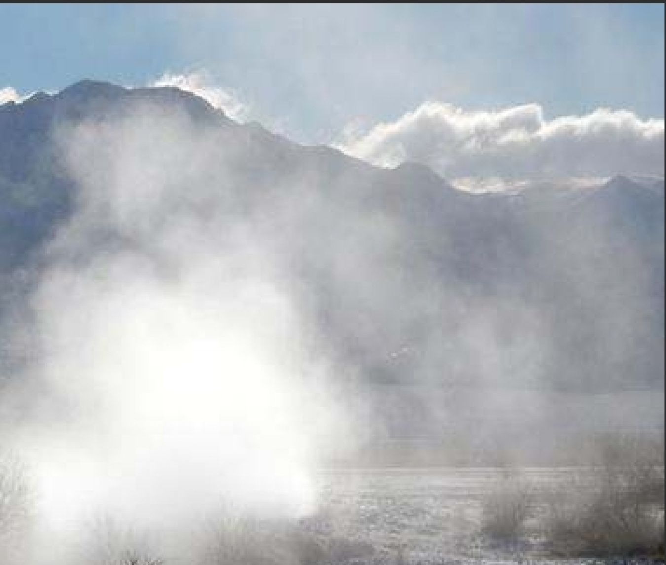 Photo of the steam eminating from a geyser in the foreground with a mountain range, clouds, and blue sky in the background.
