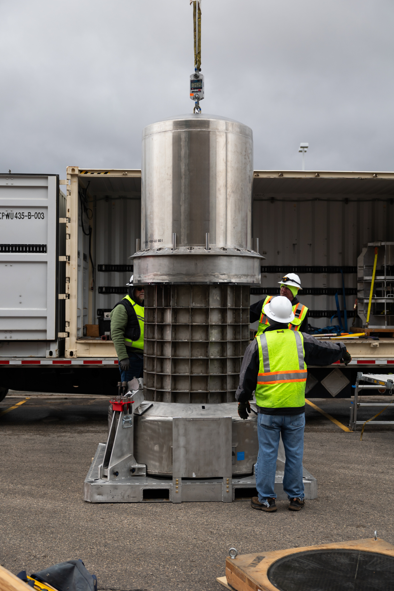 Three men help guide a crane-held transportaiton cask as part of the irradiator removal