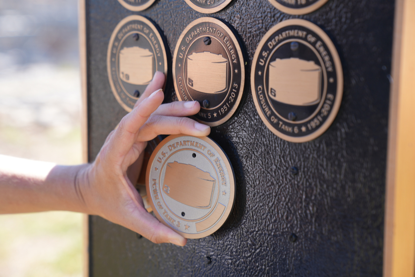 An up-close shot of a Caucasian hand placing a gold and grey circular plaque to a black wall with other plaques on it above