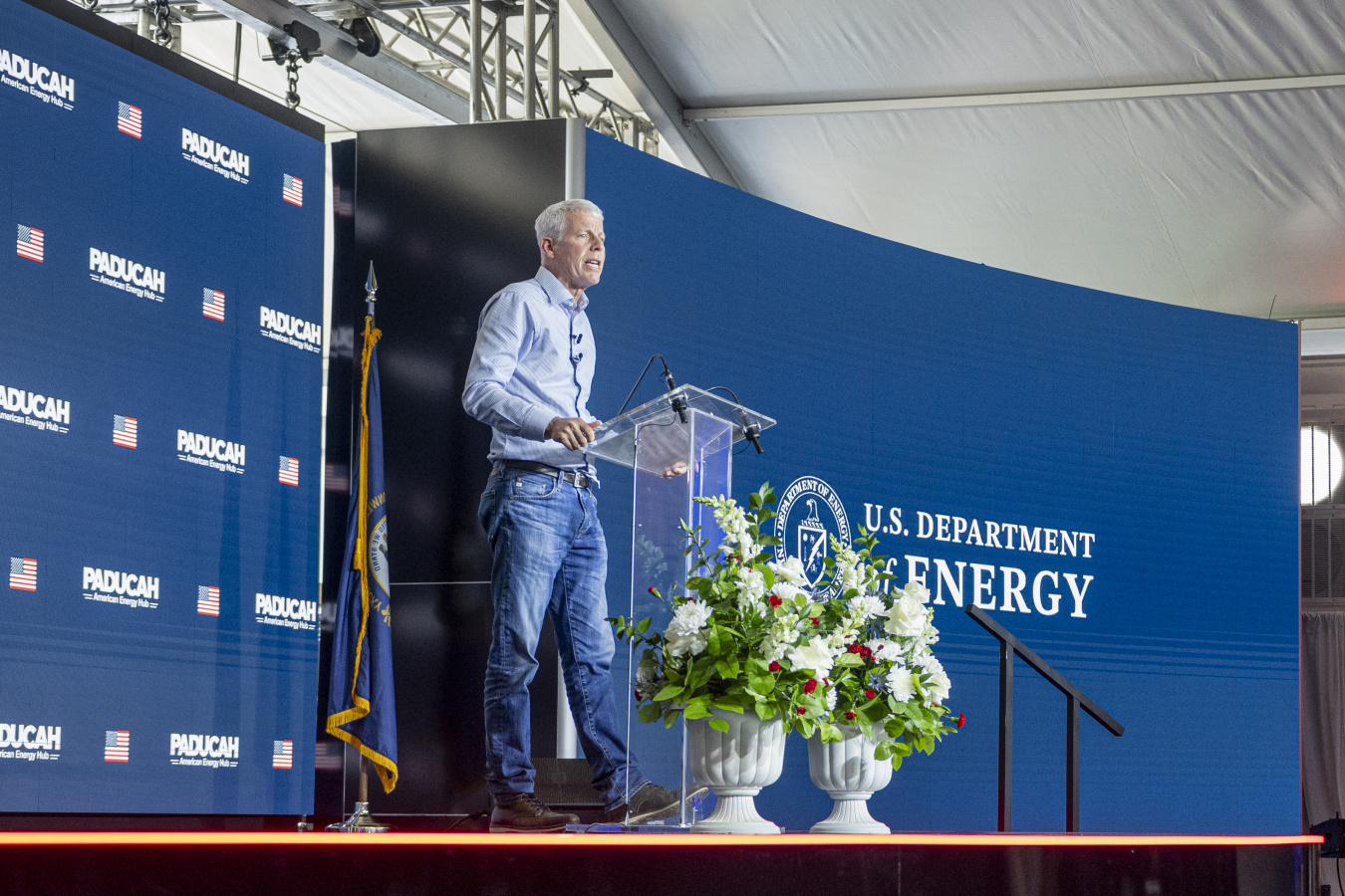 The US Department of Energy Secretary Chris Wright standing on a stage behind a clear podium with a blue backdrop behind him with the words Paducah Site and US Department of Energy written on the backdrops