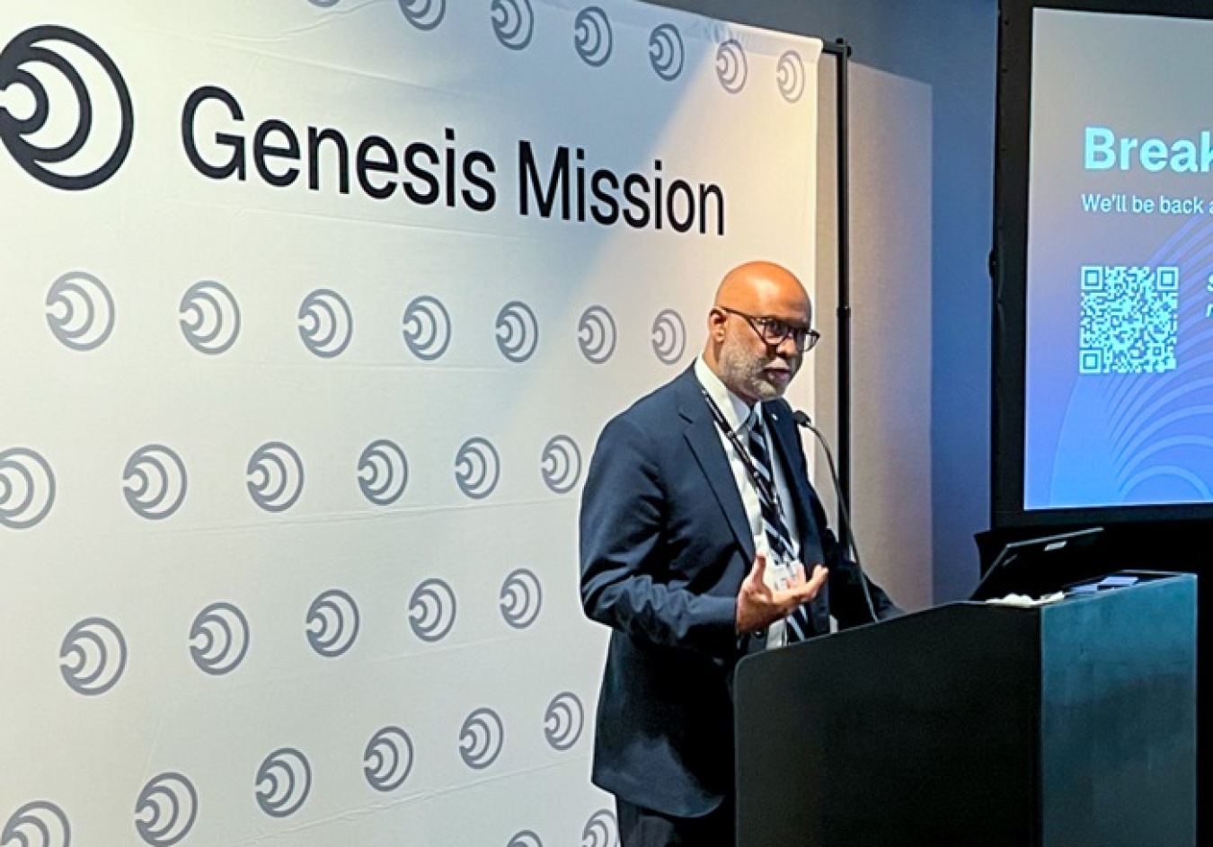 A man in a suit speaking at a podium with a presentation to his right and a white background with the words "Genesis Mission" written at the top 