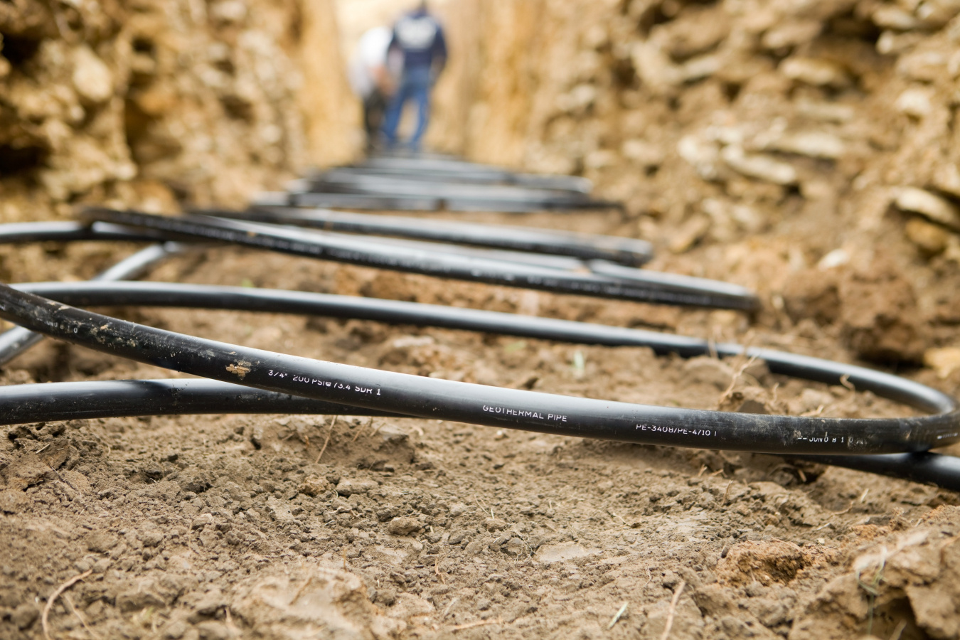 Close-up photo of black coils of piping in a trench with two figures out of focus in the background.