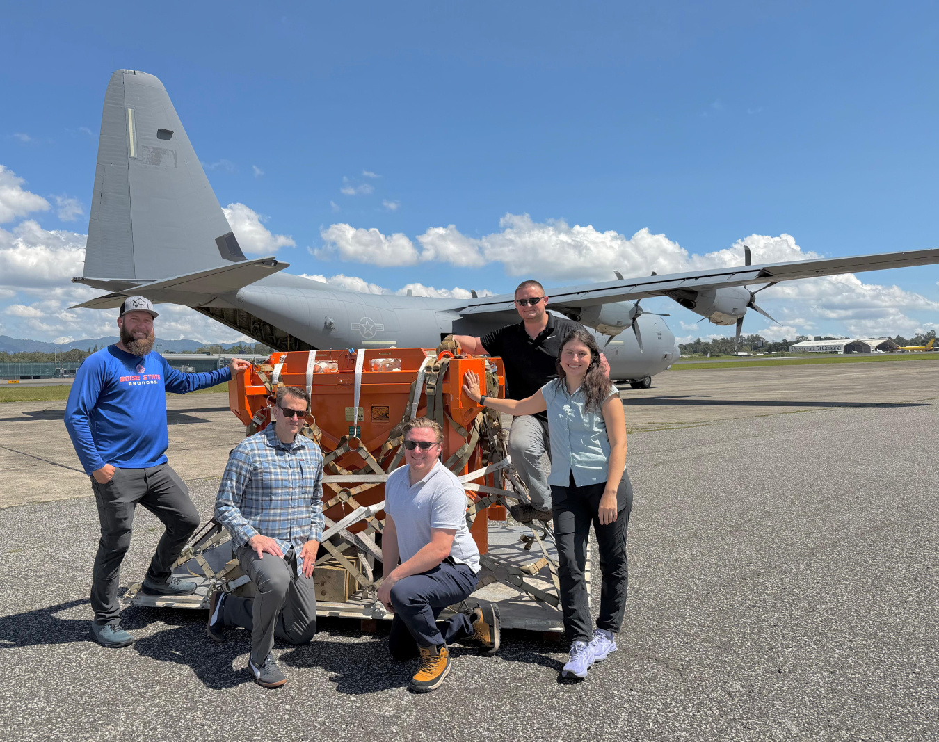 This photo shows five individuals standing around a crate. In the background there is a military transport plane.